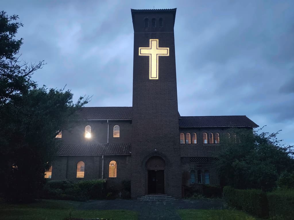 St Oswald’s Church at dusk. A glowing golden cross is projected high up on the plain brick tower above the front door.